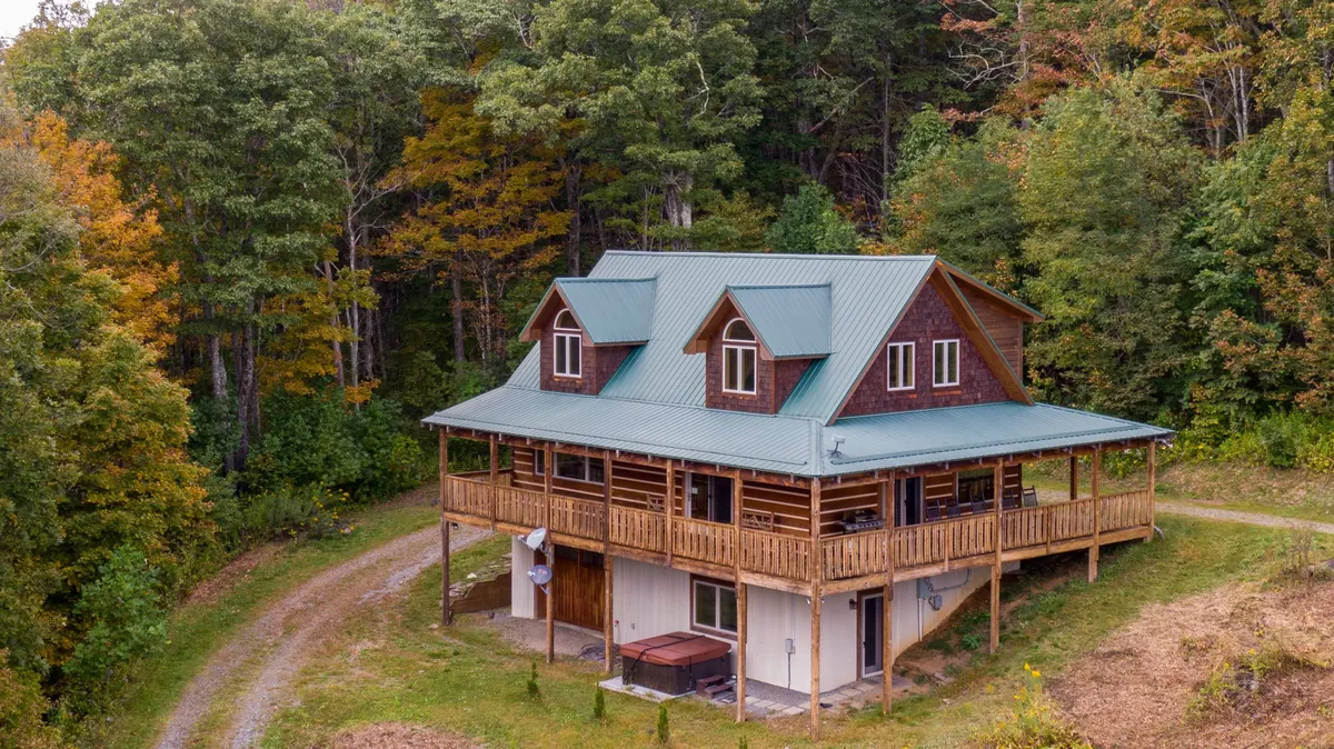 A wide view of the log cabin’s wraparound porch, green metal roof, surrounding trees, and hot tub. The image shows spaces perfect for relaxing, gathering for barbecues, and soaking in the tranquil atmosphere of the mountain setting in any season.