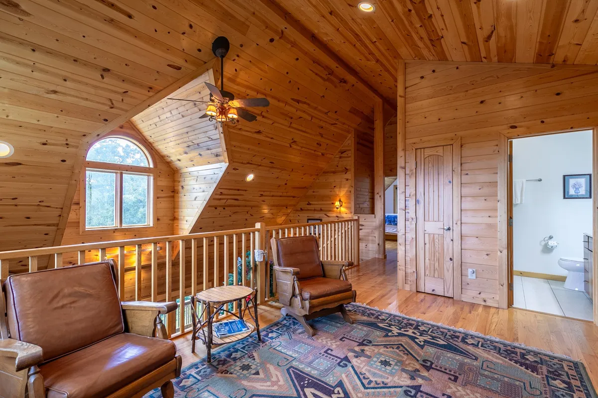 A cozy living area with a wooden ceiling, rustic furniture, and an inviting oval window that draws attention to the outdoors. Natural light streams in, and the textured rug and furniture evoke mountain character. The scene gives a glimpse of the surrounding nature through the window, connecting the inside of the cabin to the natural world outside.