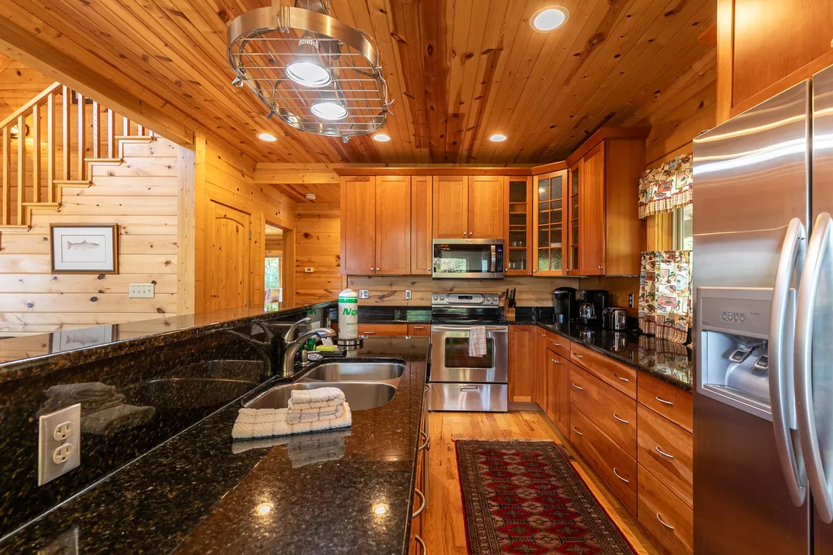 A welcoming kitchen interior featuring a gleaming granite countertop, stainless steel appliances (including a visible oven and fridge), wood cabinetry, and a patterned rug underfoot. Sunlight streams through the windows, emphasizing the warmth and inviting nature of the space.