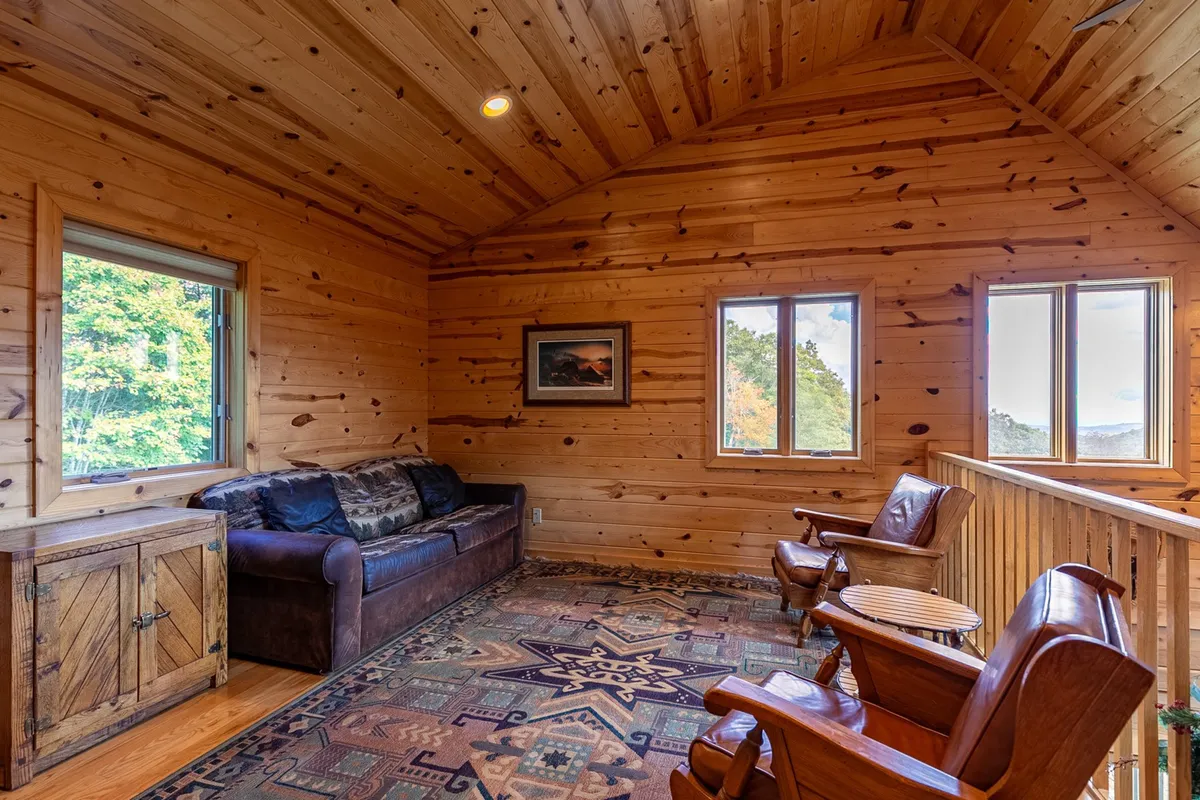 A sun-filled room with wooden furniture, large windows that frame scenic views, a textured rug, and wall artwork. The setting invites guests to unwind, socialize, and take in the natural beauty just outside—blending indoor comfort with the surrounding landscape.