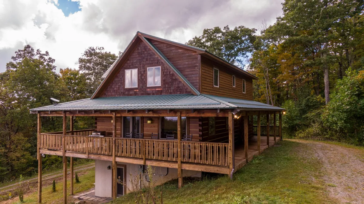 A wide view of the cabin’s inviting wooden porch, with a sloped roof and deck railing, set against a backdrop of lush trees. The space suggests outdoor gatherings, lazy afternoons, and is evocative of the area’s peaceful, scenic atmosphere.