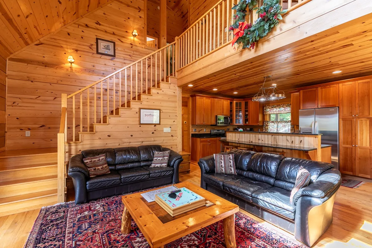A welcoming living space featuring leather sofas clustered around a wooden coffee table, with a rustic staircase and a glimpse into the open kitchen area. The room exudes warmth, with a central fireplace offering a cozy focal point, perfect for unwinding after outdoor adventures.