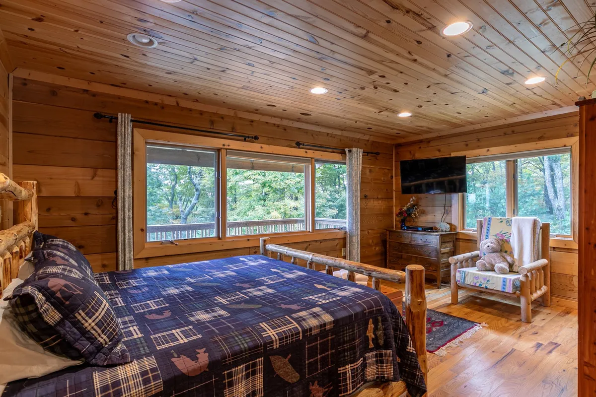A serene king bedroom featuring a log-frame bed with plush plaid bedding, large windows letting in natural light, and wooden furnishings that exude rustic charm. The space feels inviting and restful, perfect for unwinding after a day outdoors.