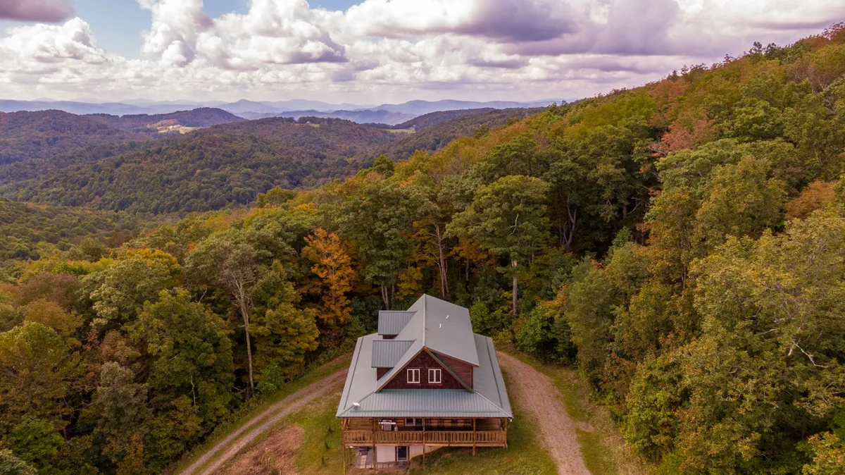 The image showcases a classic cedar log cabin nestled among tall, lush trees with expansive mountain views in the background. A wide porch runs along the front of the cabin, positioned perfectly for watching a breathtaking sunrise or unwinding under the stars. The tranquil setting highlights the natural beauty and peacefulness of the Boone, North Carolina mountains.