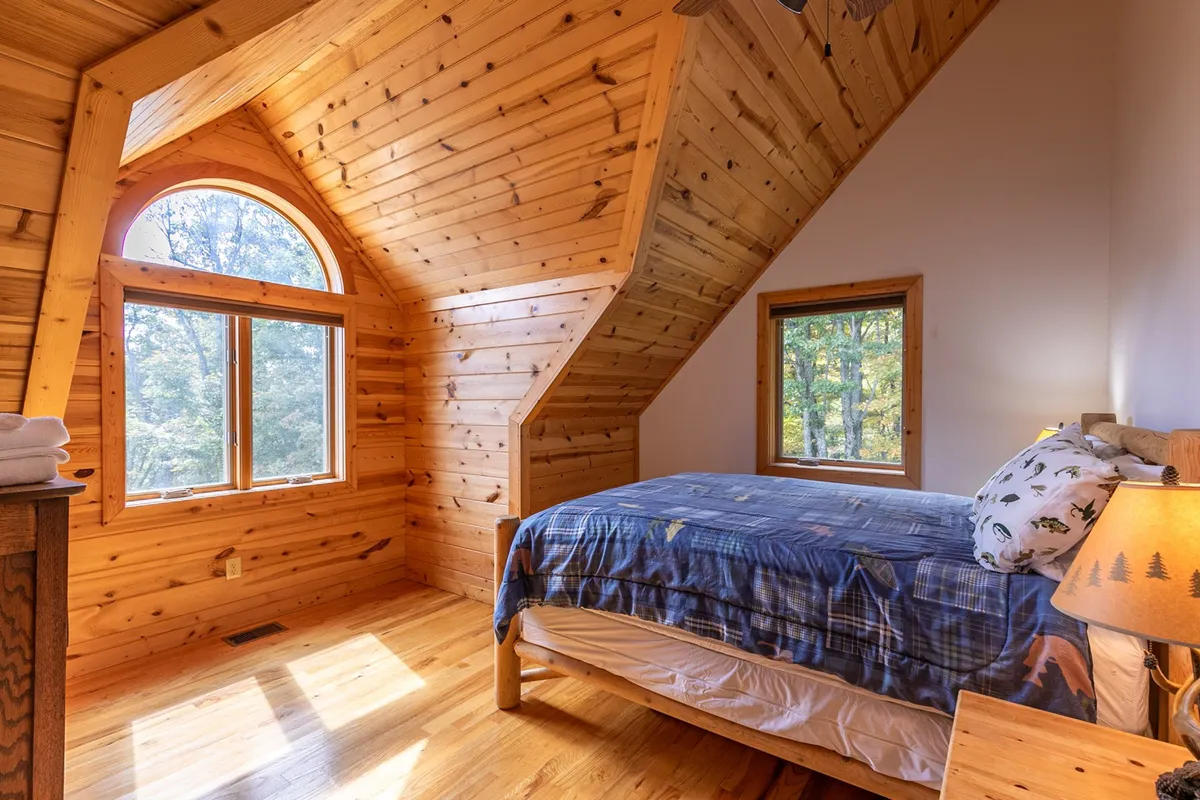 A tranquil bedroom with wooden walls, large windows that frame the view of nature outside, and soft, dappled light suggesting a peaceful cabin morning. The setting feels immersed in the landscape, reinforcing a sense of retreat and natural escape.