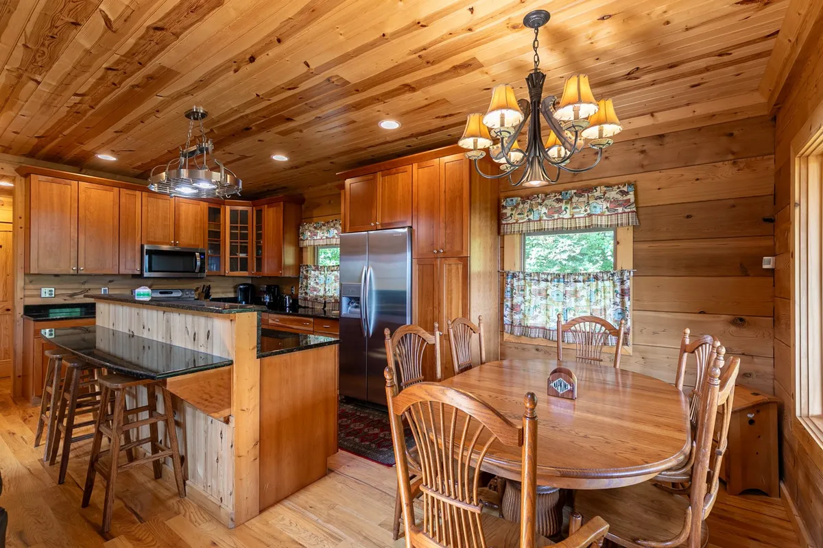 A cozy open-plan dining and kitchen area, with custom wooden cabinetry, a spacious dining table set for a group, a striking chandelier overhead, and a central kitchen island. The space invites both cooking and communal dining, with the kitchen and dining areas seamlessly integrated.