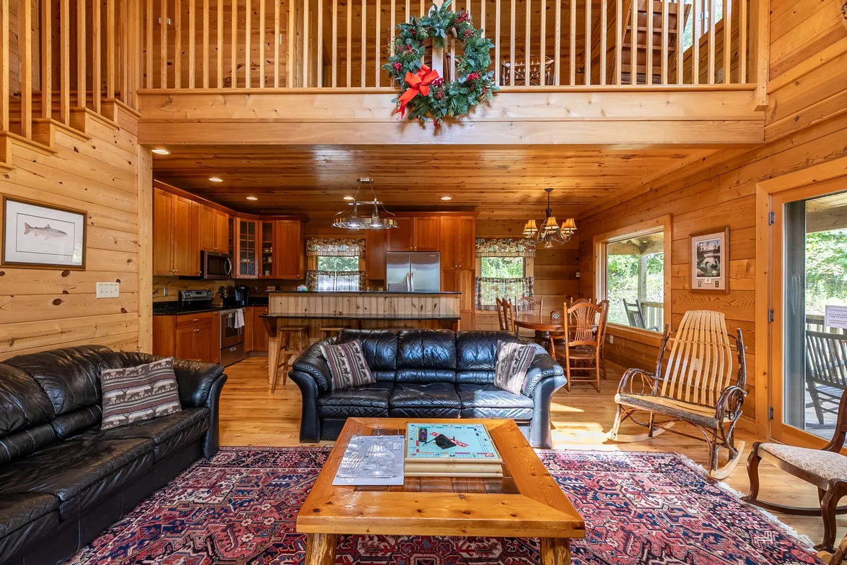 The living area showcases black leather sofas arranged for group gatherings, a large wooden dining table for shared meals, and a cozy rug. Warm lighting and a decorative wreath on the railing enhance the welcoming, social atmosphere—ideal for game nights or relaxing after a day exploring Boone.