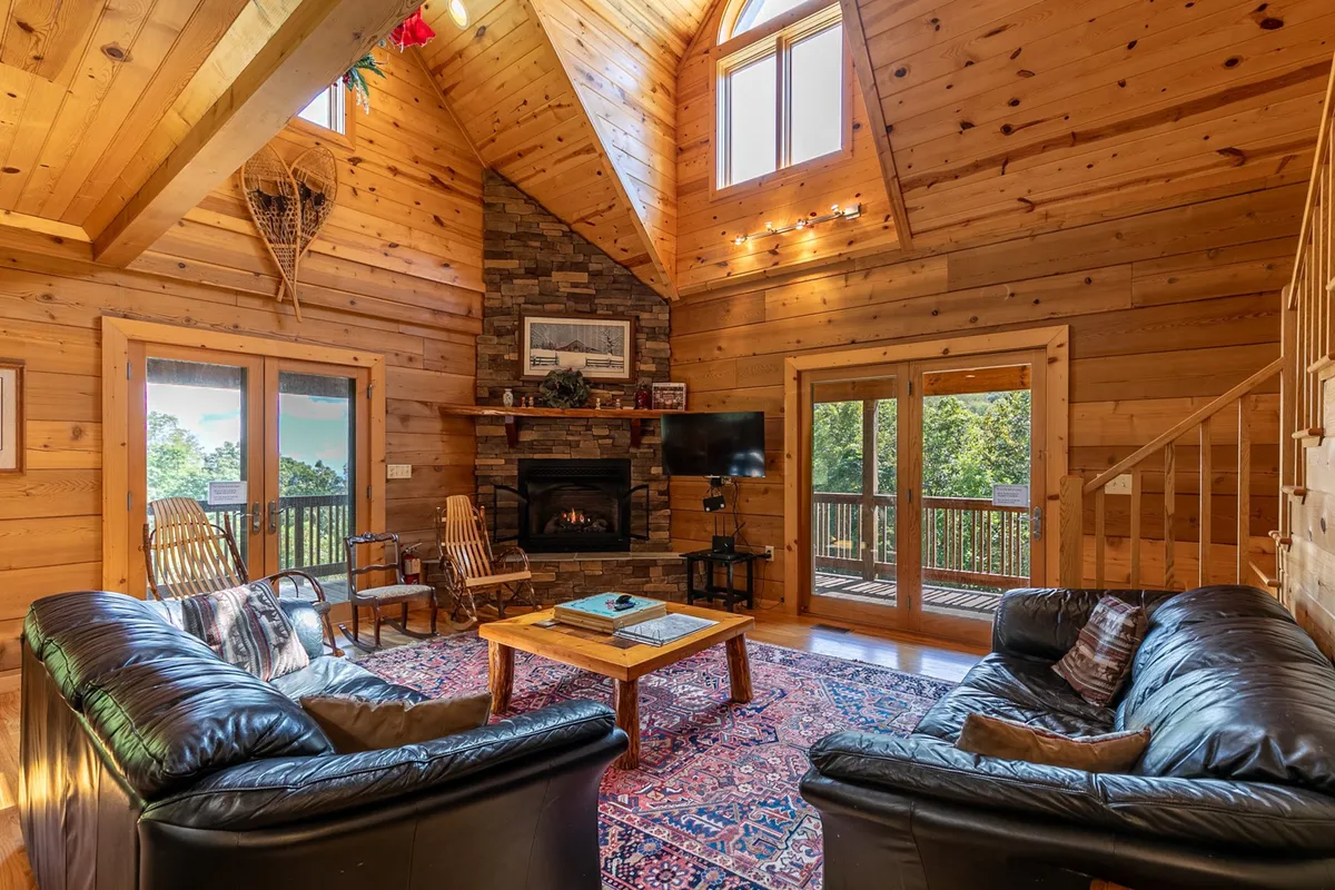 A welcoming living room anchored by a stone gas fireplace, surrounded by large windows that frame mountain views. Plush leather sofas and a wooden coffee table create a comfortable gathering area beneath a warm wooden ceiling.